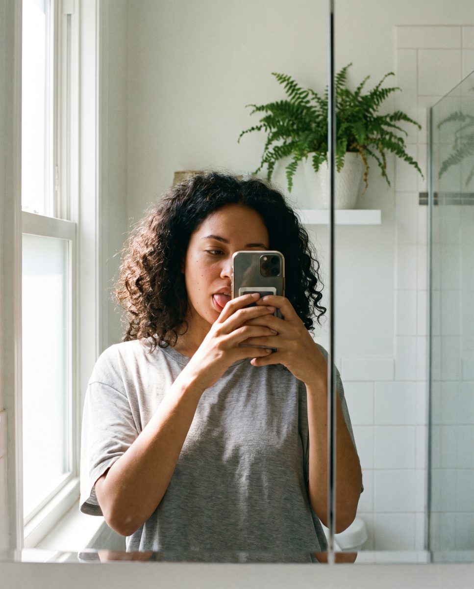 A woman shows her tongue to her phone in the bathroom mirror